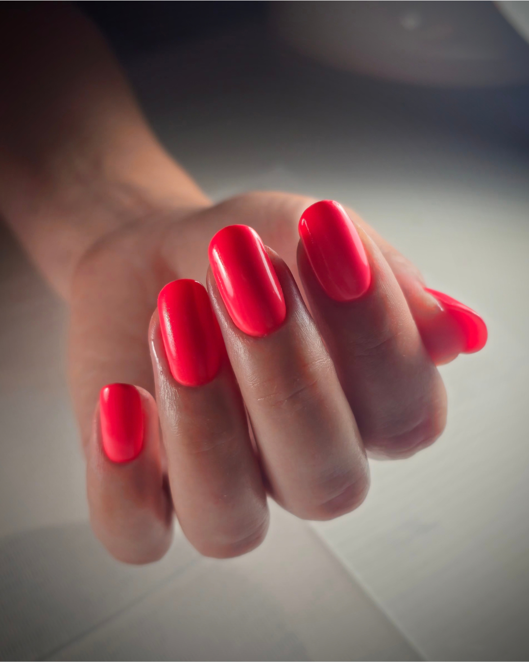 A close-up of a hand with neatly manicured nails painted in Coral, a bright, glossy neon pink-red color, set against a soft, blurred gray background. Featuring Glitch, a Phenomenail™ M.E.T.A Pro PIGM-A professional gel polish