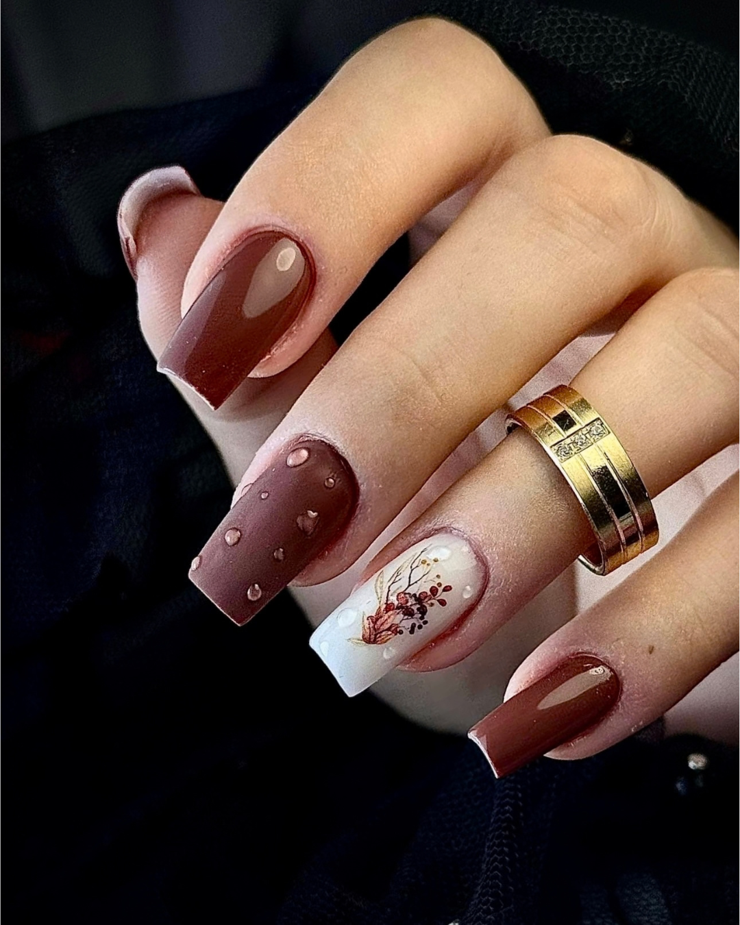 A close-up of a hand showcasing a modern coffin-shaped nail art set. The nails feature a mix of glossy dark brown, a brown base with small heart details, a white base with a delicate floral design, and a glossy dark brown accent. featuring Leather Armor chocolate-brown gel polish from the Phenomenail™ M.E.T.A Pro PIGM-A range