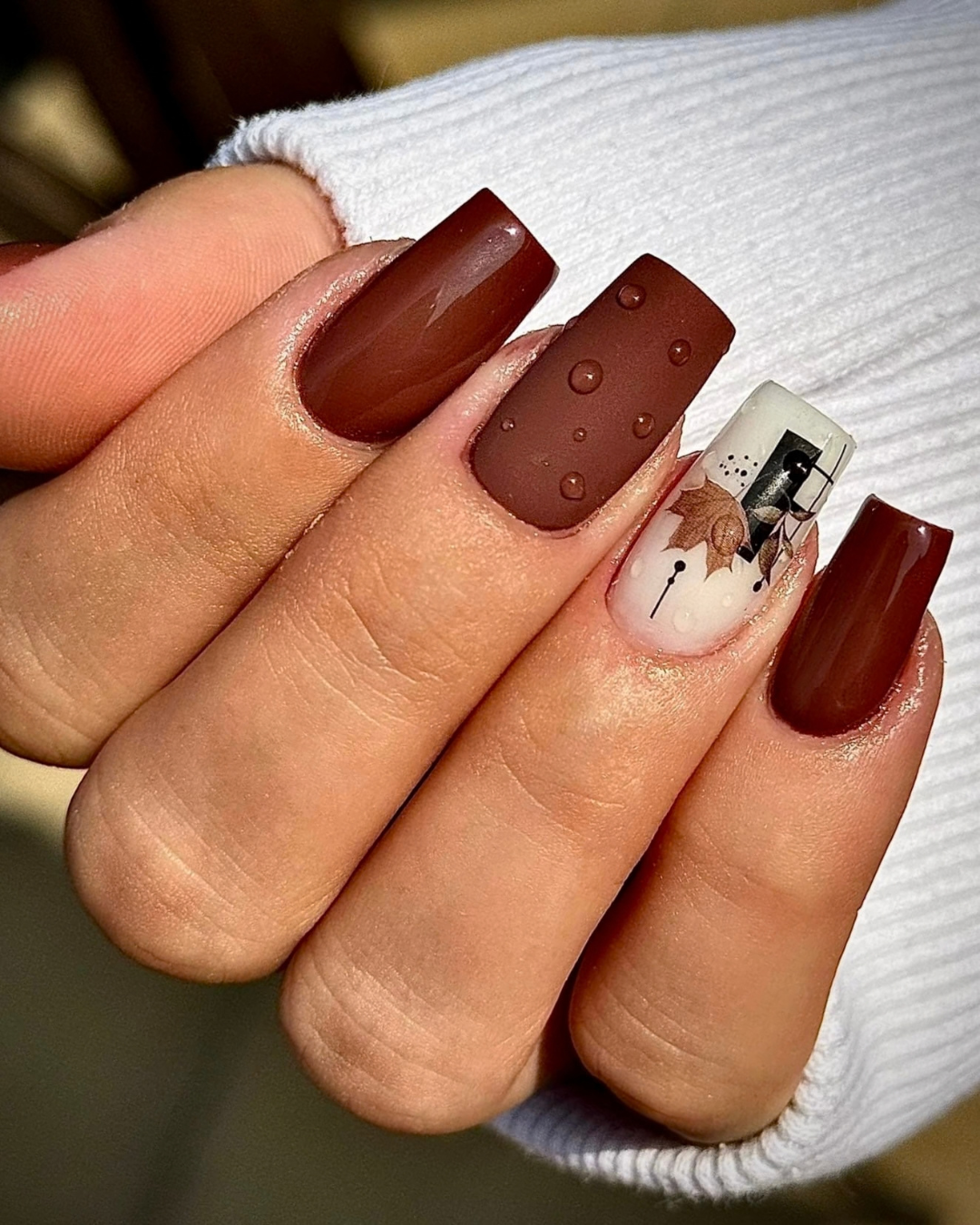 A close-up of a hand showcasing a manicure with four long, square-shaped nails. Three nails are a glossy dark brown, and the middle nail features a matte brown dotted pattern. Featuring Leather Armor chocolate-brown gel polish from the Phenomenail™ M.E.T.A Pro PIGM-A range