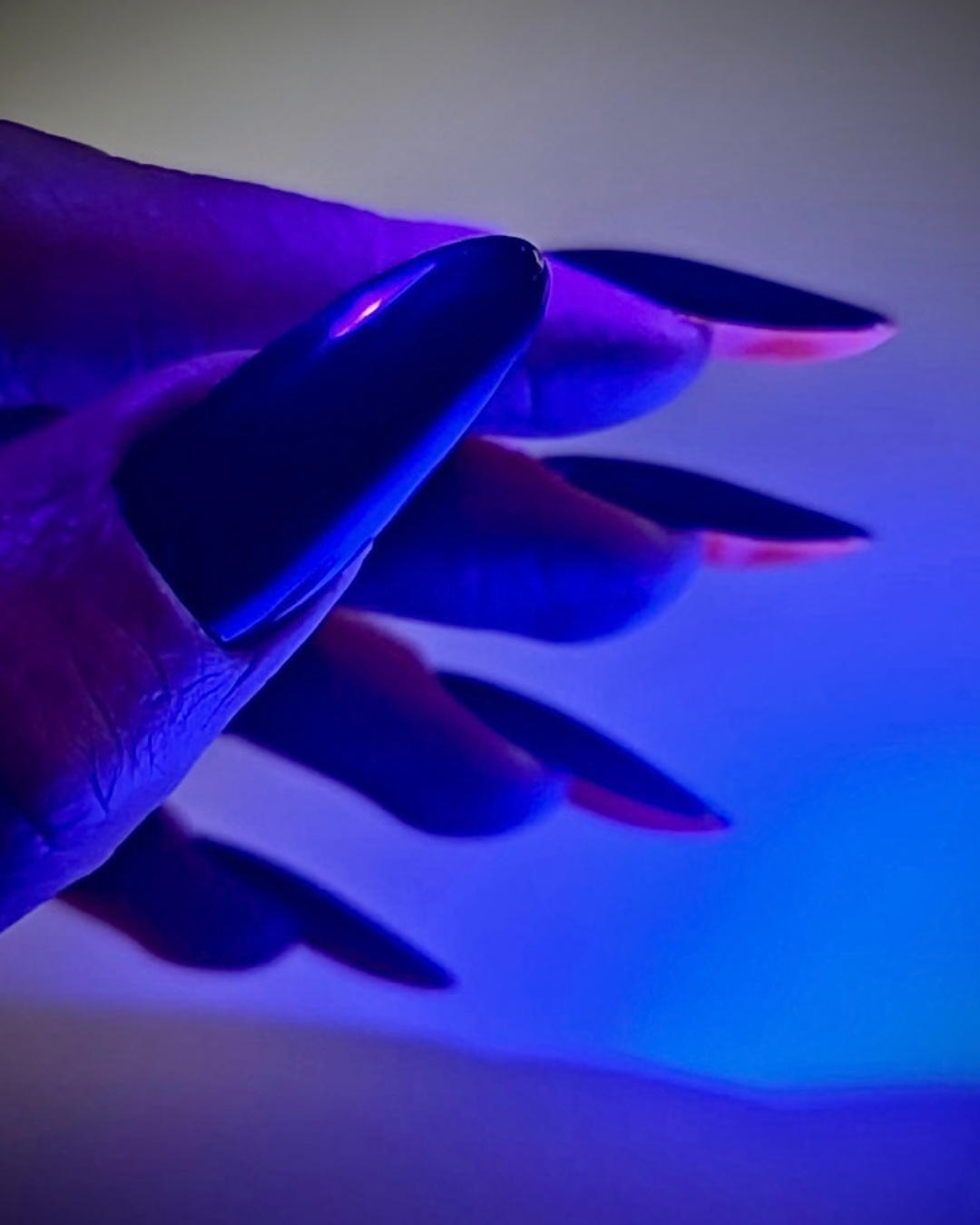 A close-up of a hand with long, glossy black manicured nails under blue and purple UV light, showing a pinkish cured edge on some nails.