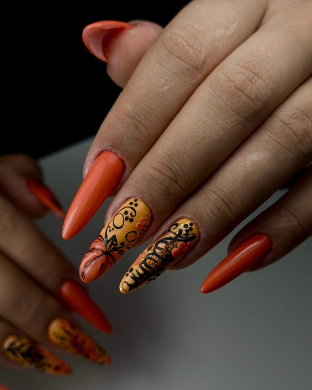 Close-up of a hand with long stiletto-shaped nails painted in orange and yellow, with one nail featuring a black floral pattern and the others showing a glossy orange finish. Featuring Gold Coin, a Phenomenail™ M.E.T.A Pro PIGM-A professional gel polish