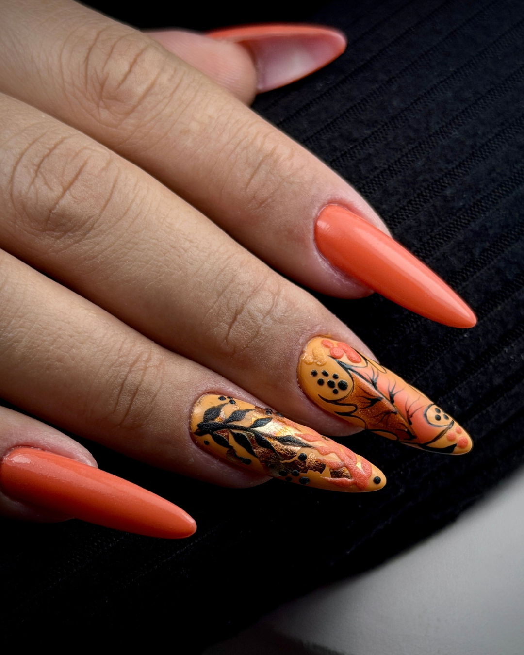 A close-up of a hand with long almond-shaped nails, featuring a solid coral-orange manicure on three nails and a detailed Halloween-themed nail design on the ring finger with orange, black, and cream floral and pumpkin motifs. Using Gold Coin, a Phenomenail™ M.E.T.A Pro PIGM-A professional gel polish