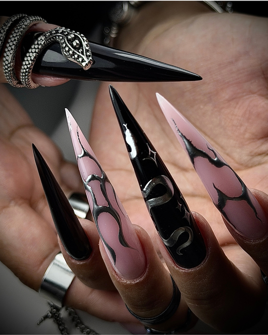 A close-up of hands showcasing long, sharp stiletto-style nails. The nails features Phenomenail™ M.E.T.A Pro Chance black gel and pale pink ombre design with silver snake graphics and black swirl patterns.