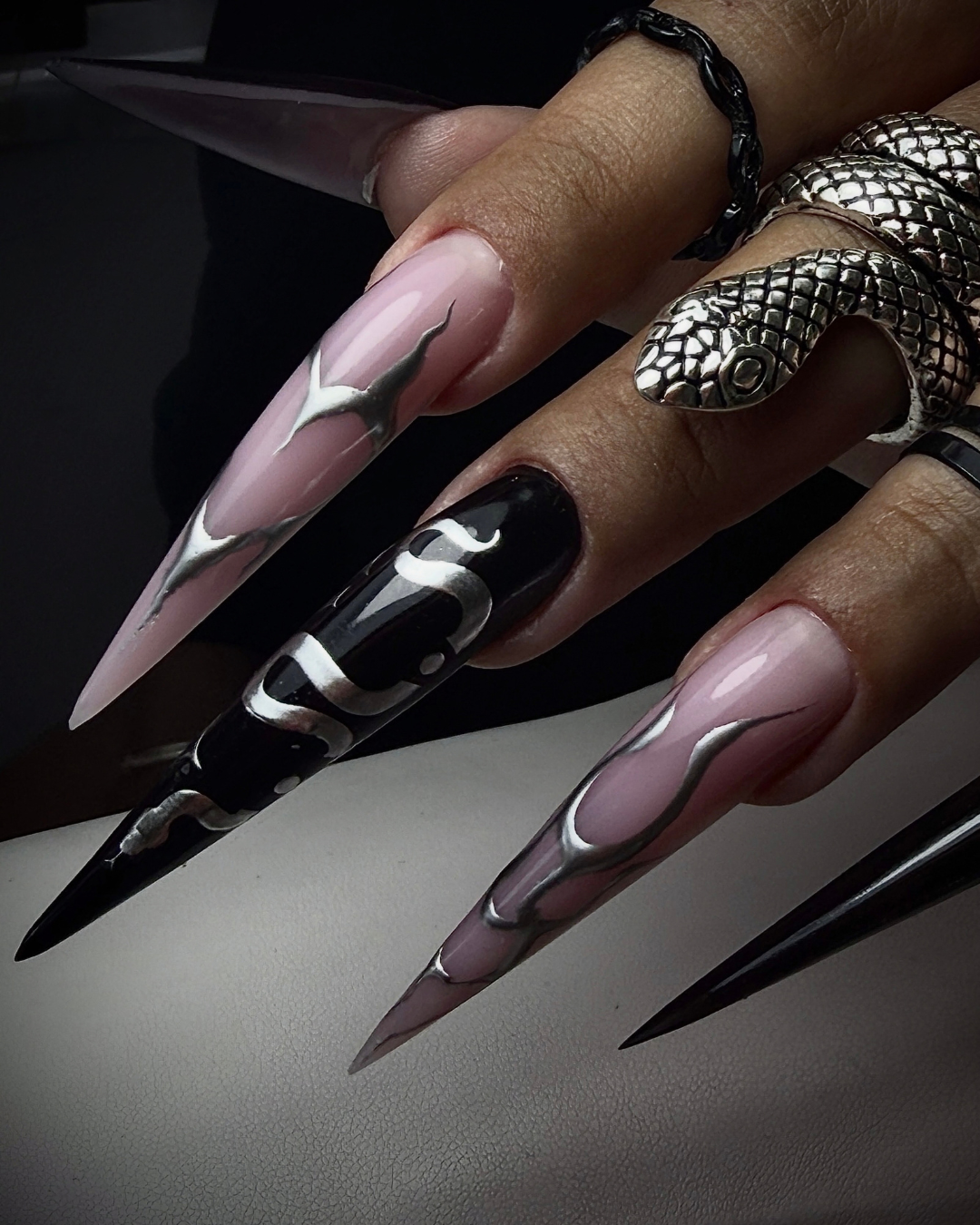 A close-up of a hand with long, sharp stiletto-style press-on nails. The nails feature a glossy pale pink base with silver metallic flame-like swirls on three nails, and a solid glossy black base with silver metallic swirls on one nail. Featuring Chance from  PIGM-A Gel Polish collection