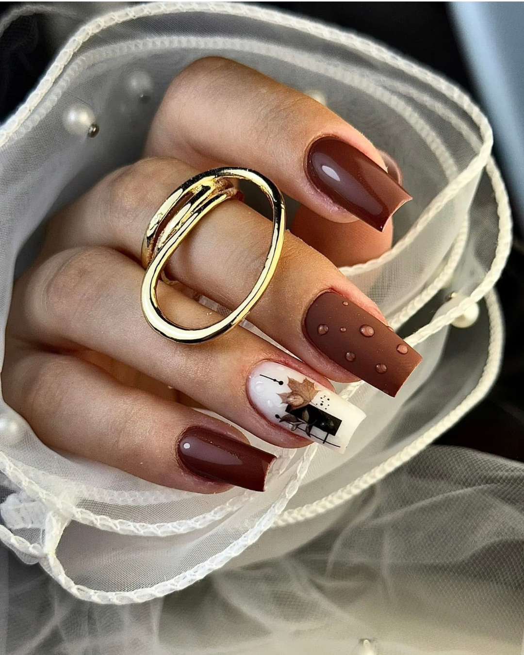 A close-up of a hand with long square nails styled in a fall theme, featuring two glossy dark brown nails, one medium brown nail with water droplets, and one white nail with a black leaf design.  Featuring Leather Armor chocolate-brown gel polish from the Phenomenail™ M.E.T.A Pro PIGM-A range
