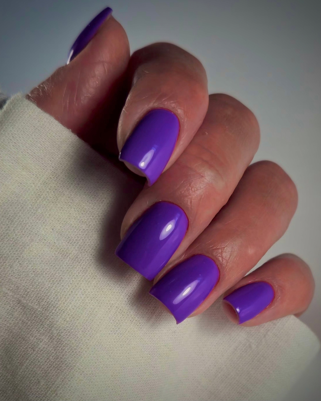 A close-up of a hand with glossy, vibrant purple manicured nails resting on a textured off-white fabric sleeve, set against a neutral gray background.