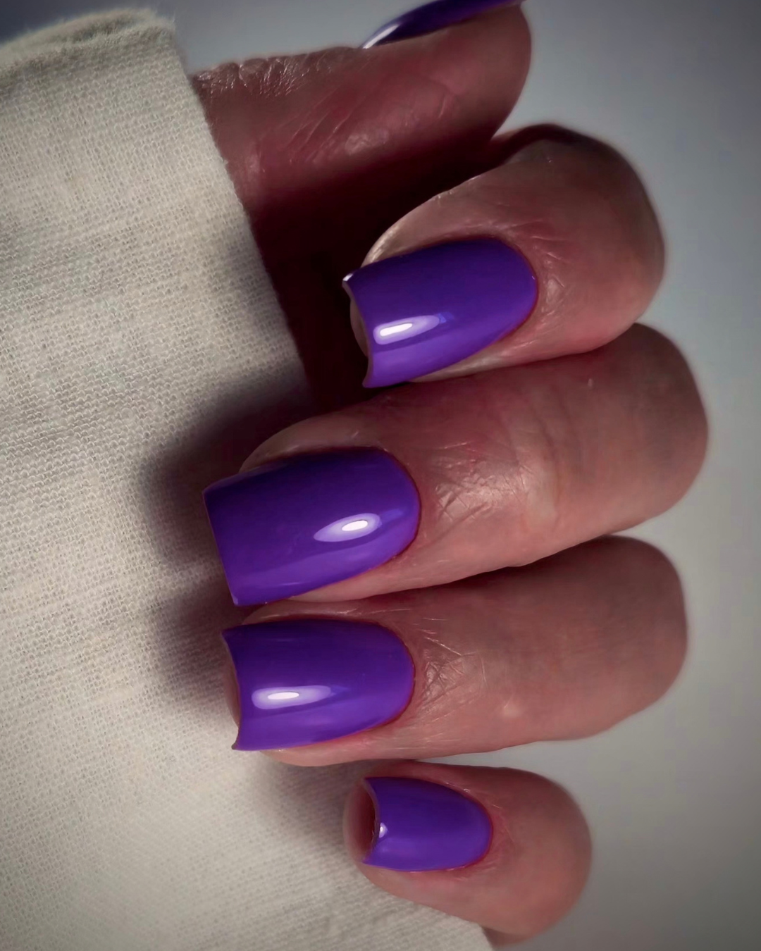 A close-up of a hand with Phenomenail™ M.E.T.A PWNED purple manicured nails, resting against a light-colored textured fabric.